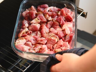 Woman hand putting a glass baking tray with pieces of raw pork meat with spice in to an oven.