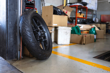 Black motorcycle alloy wheel and tire leaning against a wall in a busy auto repair workshop.