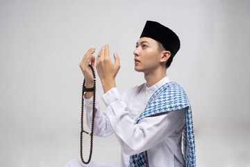 Young Asian man praying with tasbih, wearing white traditional shirt and black cap, scarf draped over shoulder, hands raised