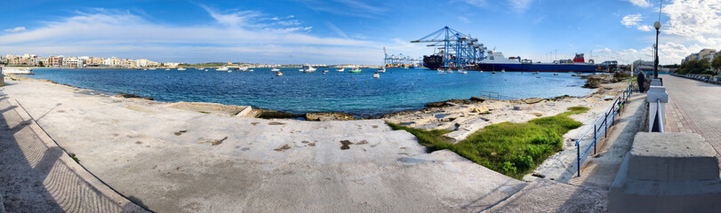 Panoramic view of Birzebbuga harbor with promenade, cargo ships, boats and houses under blue sky