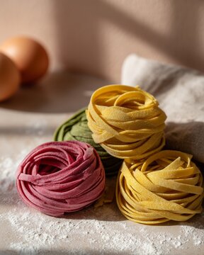 Assorted naturally dyed pasta nests in beet pink, spinach green, and golden yellow arranged on a light surface with eggs and flour in the background