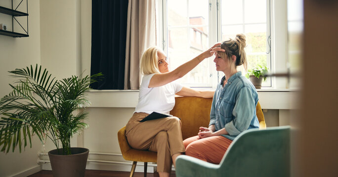 A coach practicing a touch therapy session with a client. She has placed her hand on the client&rsquo;s forehead, and the client has their eyes closed in receptivity. They are sitting on a yellow sofa