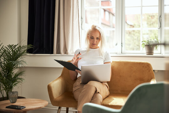 A smiling psychologist conducting online therapy, sitting on a yellow sofa in a well-lit room.