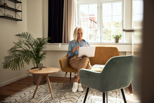 A smiling psychologist conducting online therapy, sitting on a yellow sofa in a well-lit room.