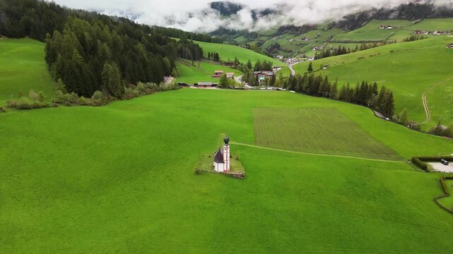 4K aerial of a small alpine chapel standing alone in green meadows and rolling hills, Val di Funes, Dolomites, South Tyrol, Italy. Low clouds and spring colors. Filmed May 6, 2025.