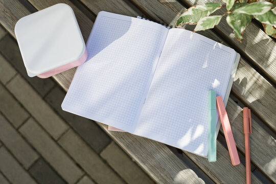 Overhead view of a blank notebook with checked paper, pens and a lunch box on a park bench in the sunlight