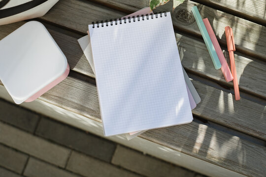 Overhead view of a blank notebook with checked paper, pens and a lunch box on a park bench in the sunlight