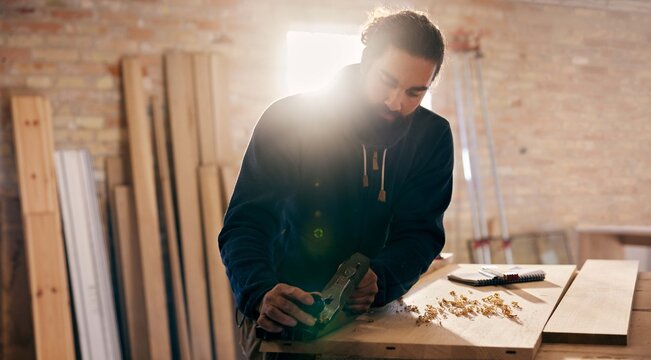 Woodworker hand planing wood at a workshop bench. Golden rays of sunlight pour in through the window behind him. Tools are placed beside him.