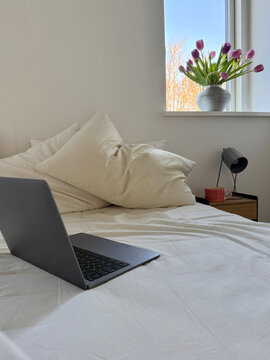 Open laptop computer on an unmade messy bed with a cup of tea on a bedside table in a bedroom