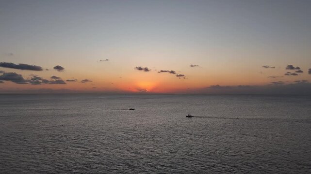 Aerial drone view of a lone fisherman rowing his small wooden boat during sunset in Vlora Bay, Albania, with soft orange skies, calm waters, and distant silhouettes of hills and clouds