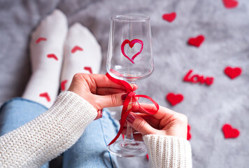 Woman's hands holding decorated wine glass with red ribbon and heart, surrounded by love decor. Romantic Valentine gifts and surprises concept. Selective focus.