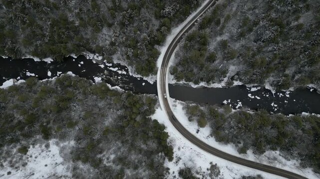 Drone top-down view of a winding road through a snow-covered forest in West Virginia with two vehicles slowly traveling through the winter landscape