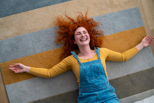 Joyful young red haired woman lying relaxed on colorful striped rug in casual denim overalls