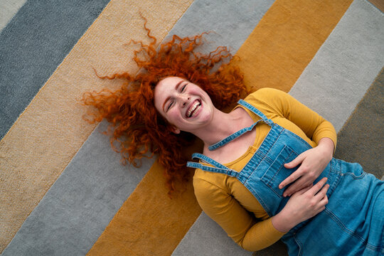 Joyful young red haired woman relaxed lying on colorful striped rug indoors smiling happily