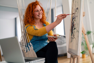 Creative young red haired woman painting abstract art in bright cozy room smiling and focused