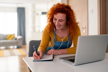 Young red haired woman smiling while writing notes using laptop at home in bright natural light