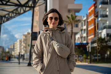 Obraz premium Young woman standing on the seaside promenade in Durrës during winter, wearing a coat. Urban coastal lifestyle concept. Cold season, city walk, and everyday life by the sea. Copy space.