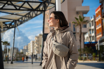 Fototapeta premium Young woman standing on the seaside promenade in Durrës during winter, wearing a coat. Urban coastal lifestyle concept. Cold season, city walk, and everyday life by the sea. Copy space.