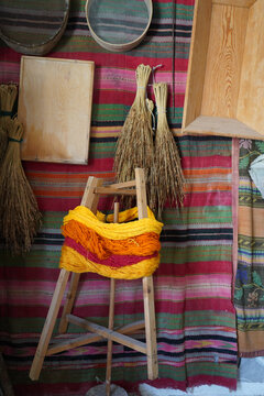 Detail of a wooden yarn winder (anemi) with yellow and red wool, set against a colorful rag rug (kourelou), wooden basins, and traditional parasires brooms.