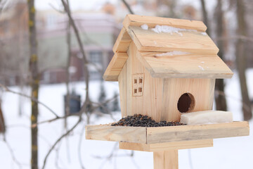 Bird feeder in winter. The feeder looks like a house with a window and a roof. A place for feeding wild birds. A beautiful wooden feeder with seeds and a piece of lard for tits. Winter © Mariia