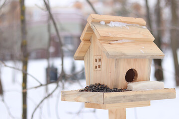 Bird feeder in winter. The feeder looks like a house with a window and a roof. A place for feeding wild birds. A beautiful wooden feeder with seeds and a piece of lard for tits. Winter © Mariia