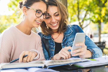 Two happy students sharing smartphone outdoors while studying