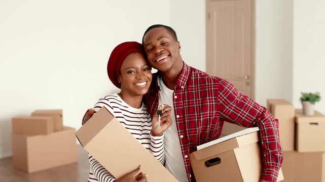 A man and woman with dark skin smile happily while holding moving boxes in a bright room. They are clearly excited about their new space and the start of a new chapter.