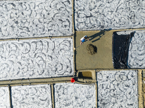 Aerial view of a solitary figure toiling amidst vast, textured salt pans under the bright sun, creating a stark contrast of light and shadow, Cox's Bazar, Chittagong Division, Bangladesh.