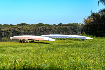 Two long canoes side by side lie upside down on the grass  on the shore of a lake.