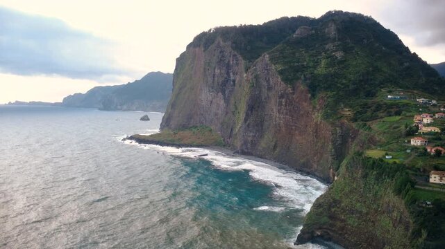 Drone descending over the high rugged cliffs of Penha d'Aguia near Faial, view towards Sao Lourenco, massive coastal rocks, Atlantic ocean waves and village houses on the edge in Madeira island.
