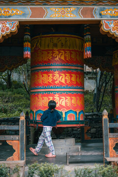 Paro, Bhutan - 16 September 2025: View of a child in patterned pants spins the vibrant prayer wheel, its ornate carvings standing out against the ancient temple's weathered wood.