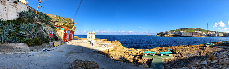 Bathing platform with umbrellas, sun benches, rocks, view of Marsalforn, sea and buildings under blue sky