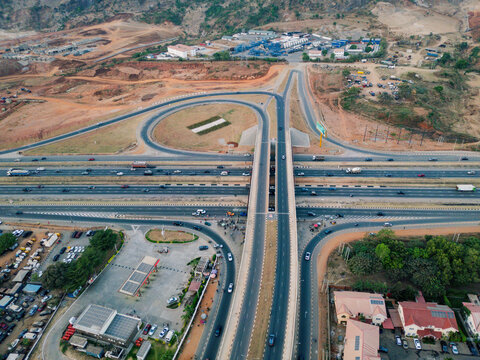 Aerial view of a complex highway interchange with multiple lanes carrying cars and trucks, framed by the reddish-brown earth and distant buildings, Abuja, Federal Capital Territory, Nigeria.