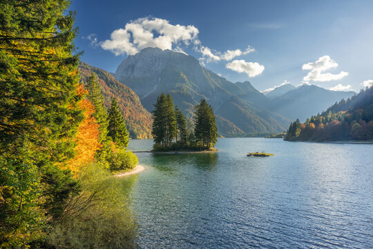 View of a tranquil lake reflects the surrounding mountains and a small, tree-covered island under a bright sky, near a colorful forest, Friuli-Venezia Giulia, Italy.