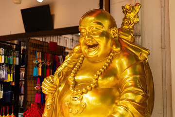 Fototapeta premium A large golden statue of a smiling figure stands in a shop filled with colorful items. Bright lights reflect off the shiny surface of the statue. This scene captures the vibrant energy of the market