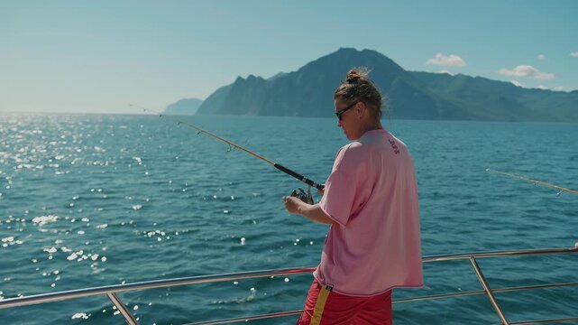 Young man with ponytail and sunglasses fishes with spinning rod from yacht during deep-sea fishing trip in Pacific Ocean. Concept of silence and pacification