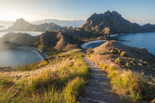 View of a rugged path leads down to pristine beaches nestled between dramatic, mountainous islands under a serene sky, West Manggarai Regency, Indonesia.