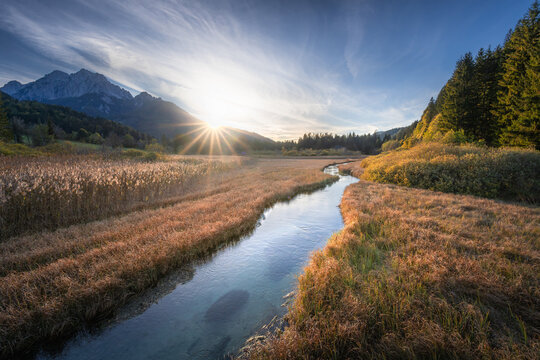 View of a serene river winding through golden fields, reflecting the sky under the gaze of distant mountains, Kranjska Gora, Jesenice, Slovenia.