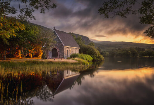 View of a stone chapel, with a dark roof reflected in the still waters of the lake, framed by trees under a fiery sunset sky, Bantry, Ireland.