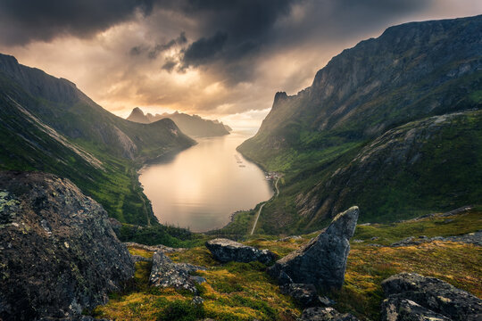 View of dramatic cliffs rise from the still waters under a foreboding sky, with foreground rocks adding texture and depth, Troms, Norway.