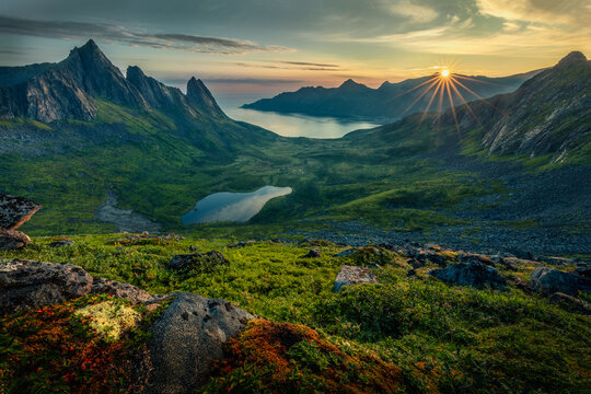 View of sunlight streams over the horizon, illuminating jagged peaks, green valleys, still lake and rocky foreground, creating a serene landscape, Troms, Norway.