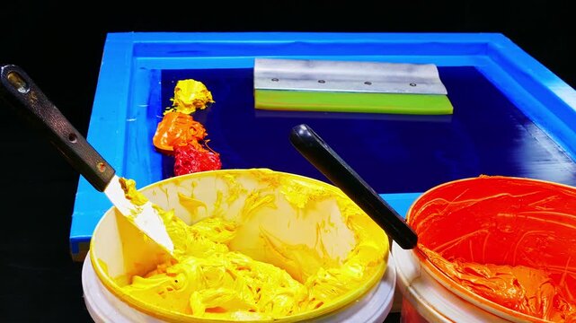 A close up at a colorful screen printing station filled with bright yellow, orange, and red inks. Spatulas rest beside a blue printing frame, capturing the energy and precision of artistic creation