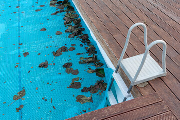 Ladder At Pool Edge With Leaves, Wooden Deck Planks Framing Metal Steps, Blue Cover And Scattered Debris, Calm Water Surface, Safety Concern For Swimmers, Area Awaiting Maintenance Inspection