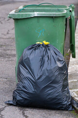 a large, tied black trash bag filled with waste, resting on the pavement in front of a green plastic wheelie bin.