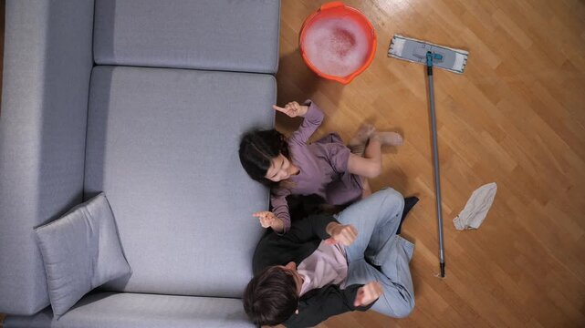 Father and young daughter take a relaxed break on a wooden living room floor beside a mop and sudsy bucket, sharing a candid moment during cleaning chores together