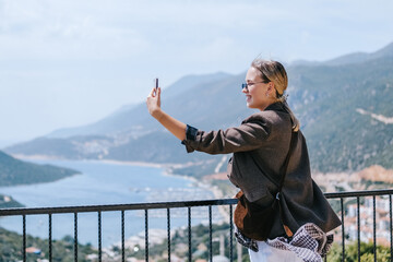 Travel blogger filming live stream on smartphone against mountain coastline, Gen Z female tourist on viewpoint, panoramic sea and hills, social media content creator, sunny outdoor vacation vibe. © shintartanya