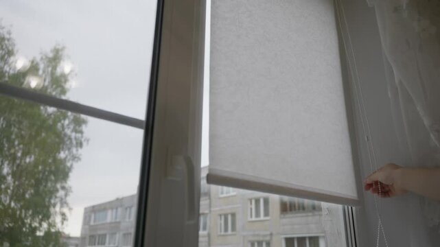 Close-up shot of a human hand pulling a cord to lower a Roman window blind. Home interior detail, light control process and everyday domestic routine inside an apartment. High quality 4k footage