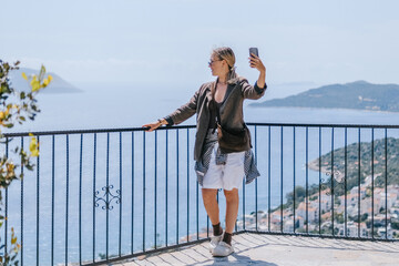 Travel blogger filming live stream on smartphone against mountain coastline, Gen Z female tourist on viewpoint, panoramic sea and hills, social media content creator, sunny outdoor vacation vibe. © shintartanya
