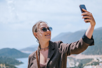 Travel blogger filming live stream on smartphone against mountain coastline, Gen Z female tourist on viewpoint, panoramic sea and hills, social media content creator, sunny outdoor vacation vibe. © shintartanya