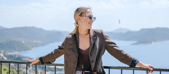 Travel blogger filming live stream on smartphone against mountain coastline, Gen Z female tourist on viewpoint, panoramic sea and hills, social media content creator, sunny outdoor vacation vibe. © shintartanya